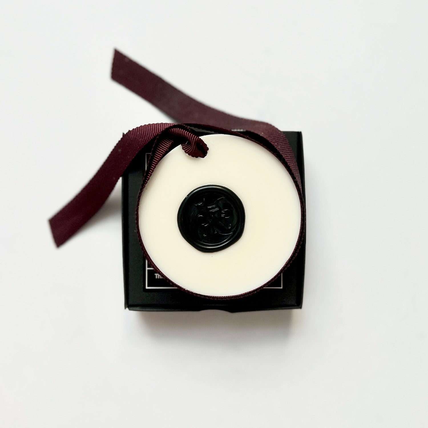 A white wax tablet with a black lid, tied with a ribbon and presented in a black box with a note card and organza bag.