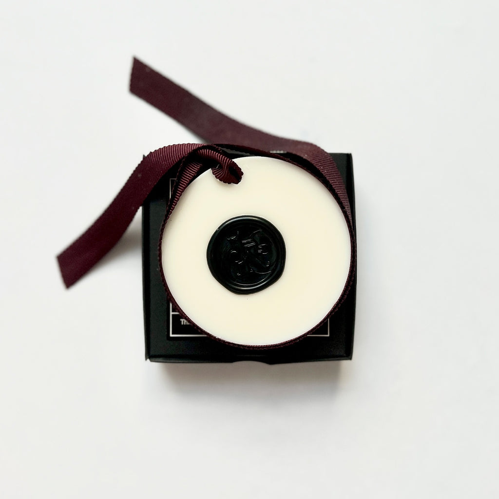 A white wax tablet with a black lid, tied with a ribbon and presented in a black box with a note card and organza bag.