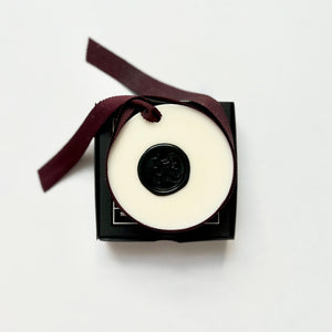 A white wax tablet with a black lid, tied with a ribbon and presented in a black box with a note card and organza bag.