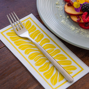 Silver fork on a yellow and white patterned napkin with a plate of fruit in the background.