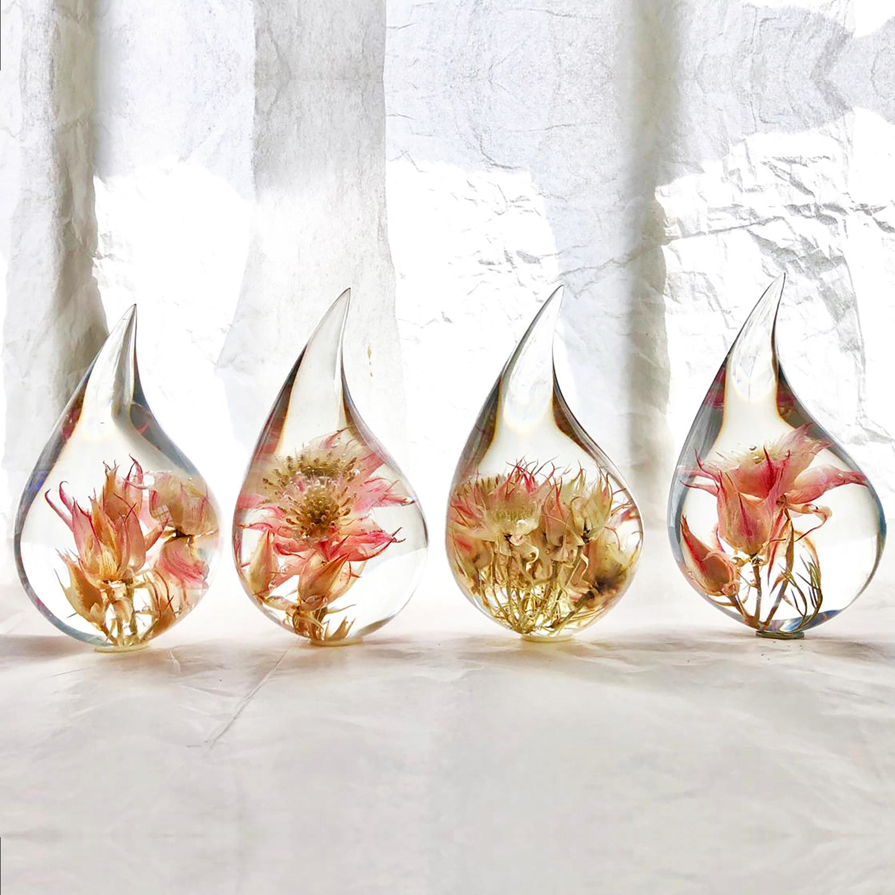 Four resin drops sculpted in a teardrop shape with pink Blushing Bride flowers embedded, positioned on a textured surface with a white background.