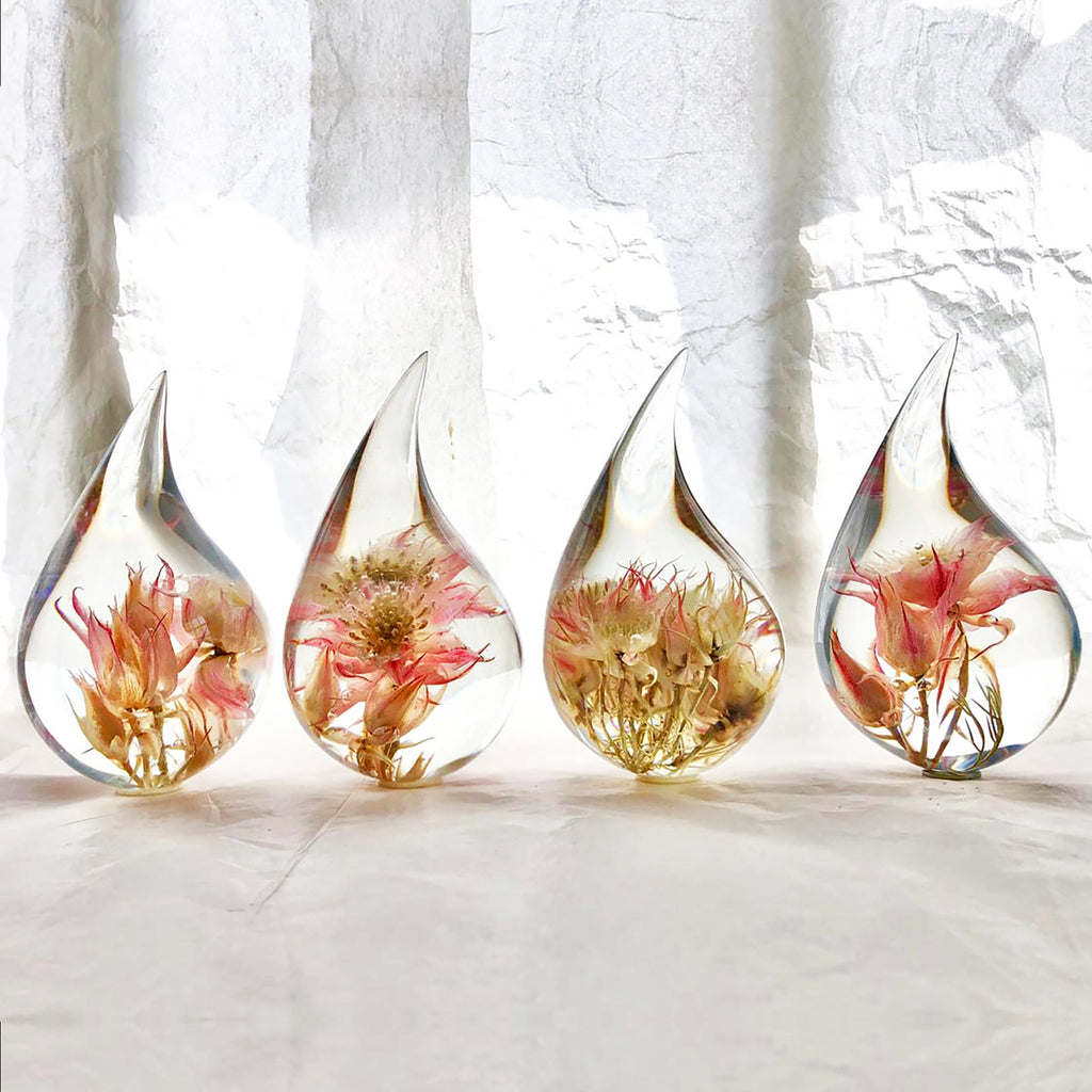Four resin drops sculpted in a teardrop shape with pink Blushing Bride flowers embedded, positioned on a textured surface with a white background.
