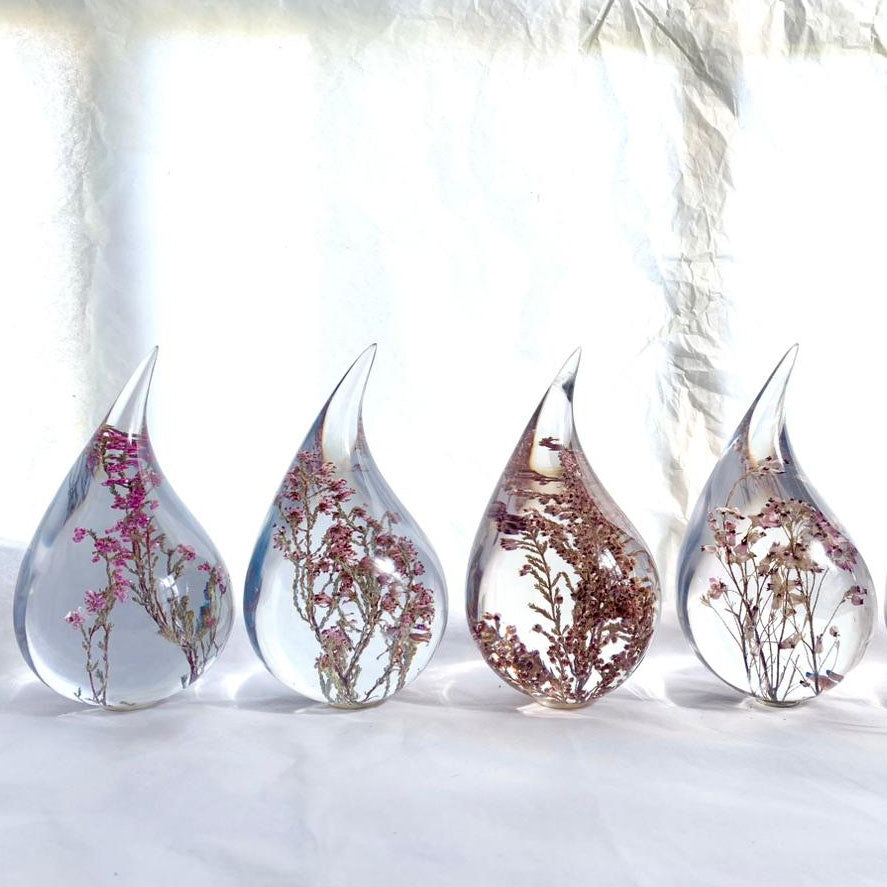 Four resin drops sculpted in a teardrop shape with purple Erica fynbos flowers embedded, positioned on a textured surface with a white background.