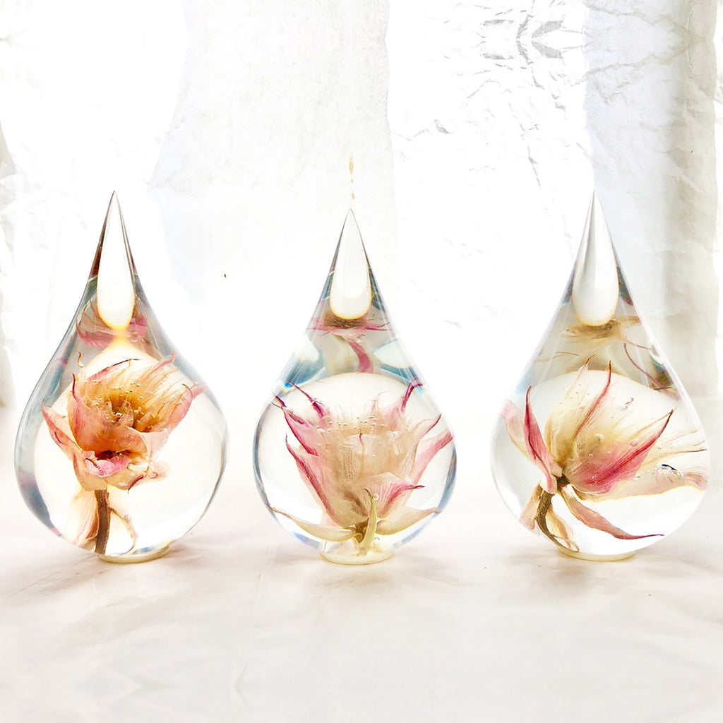 Three resin drops sculpted in a teardrop shape with Blushing Bride flowers embedded, positioned on a textured surface with a white background.
