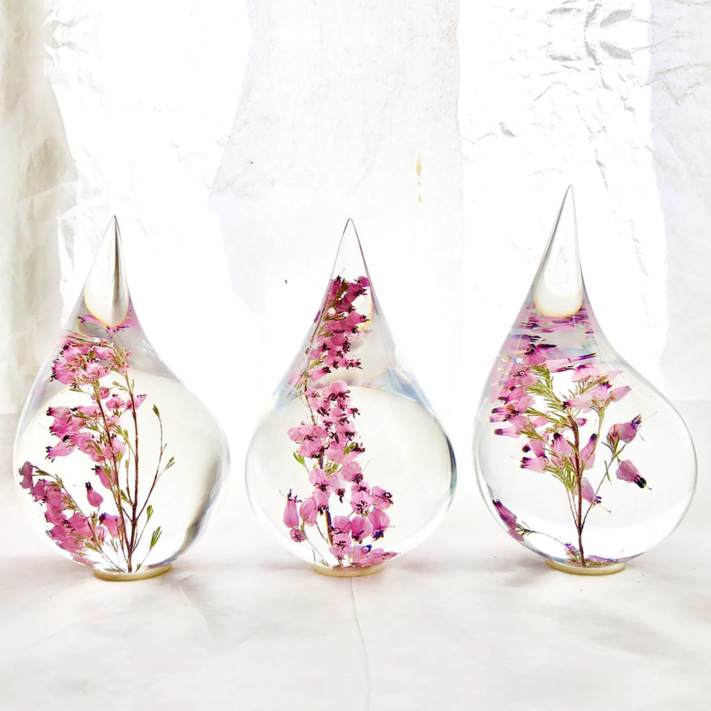 Three resin drops sculpted in a teardrop shape with purple erica flowers embedded, positioned on a textured surface with a white background.
