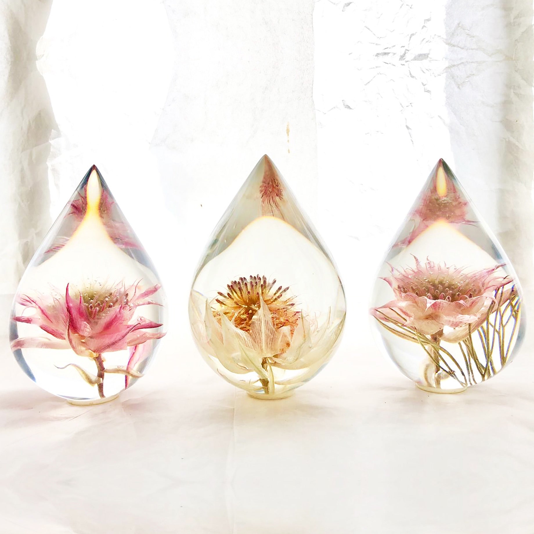 Three resin drops sculpted in a teardrop shape with blushing bride flowers embedded, positioned on a textured surface with a white background.