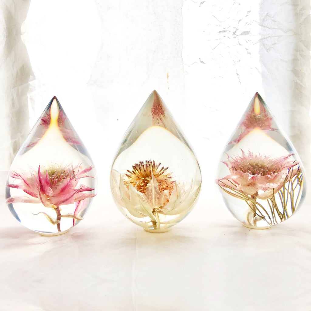 Three resin drops sculpted in a teardrop shape with blushing bride flowers embedded, positioned on a textured surface with a white background.