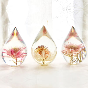 Three resin drops sculpted in a teardrop shape with blushing bride flowers embedded, positioned on a textured surface with a white background.