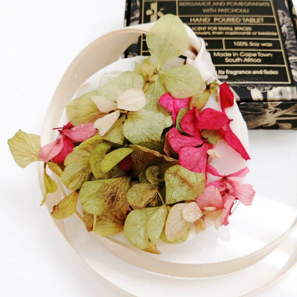 Decorative wax tablet with pink and green petals and a pink ribbon, with a box labeled 'Scented Wax Tablet Bergamot and Pomegranate with Patchouli' in the background.