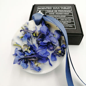 Decorative wax tablet with blue petals and a blue ribbon, with a box labeled 'Scented Wax Tablet Figue de Provence' in the background.