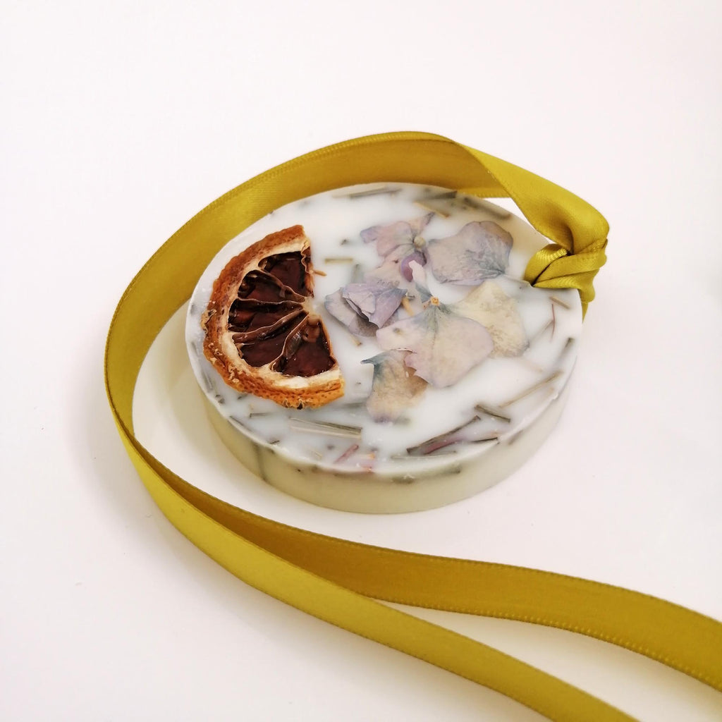 A round scented wax tablet with a slice of citrus peel and a yellow ribbon on top, placed on a white surface.
