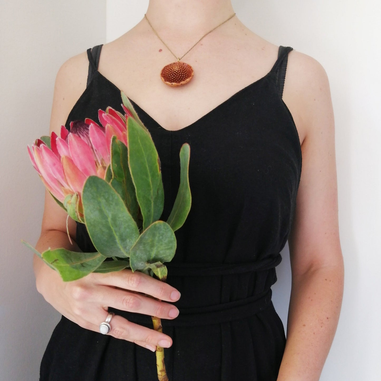 A woman holding a king protea bloom wearing a brass necklace with a protea seed pendant