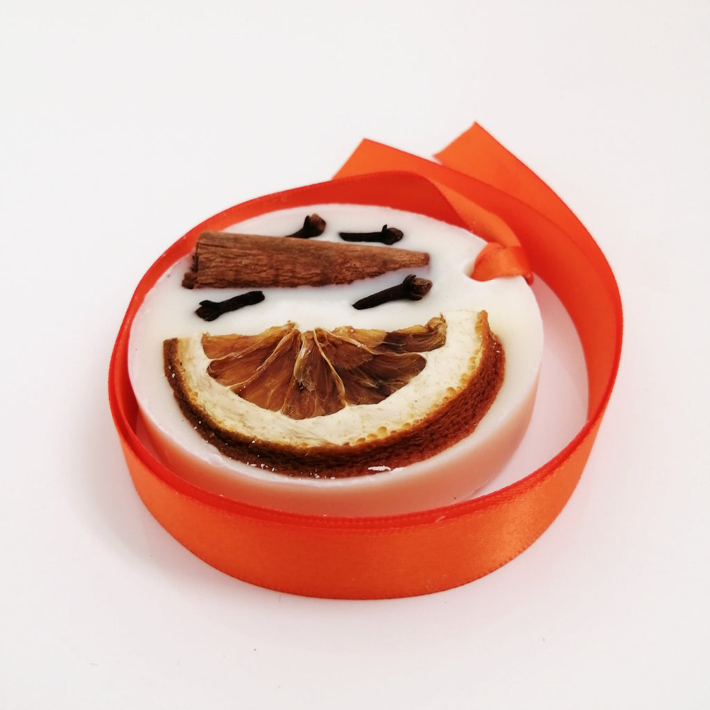 A wax tablet with a spicy orange scent, featuring a smiling face design with cinnamon sticks and orange peel slices, presented in a red ribbon package.