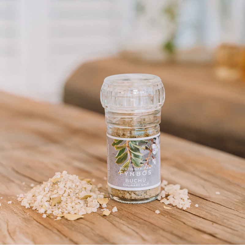 A clear glass culinary grinder labeled 'Buchu' with visible white salt crystals next to it on a wooden surface.