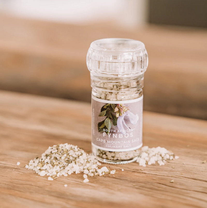 A clear glass culinary grinder labeled 'Cape Mountain Sage' with visible white salt crystals next to it on a wooden surface.