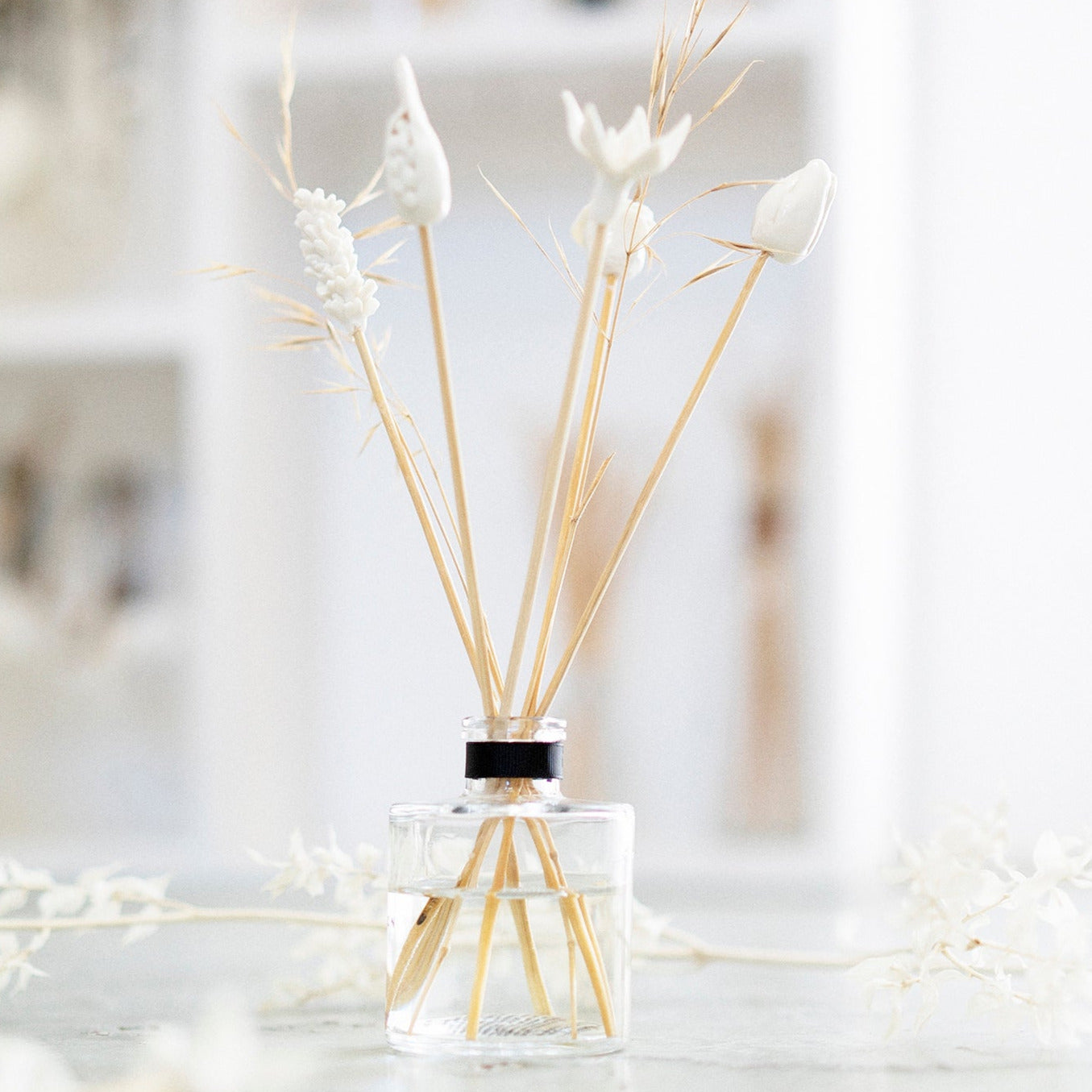 A clear glass diffuser bottle with several hand-sculpted porcelain flowers mounted on bamboo sticks, set against a softly lit background.