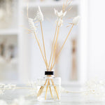 Load image into Gallery viewer, A clear glass diffuser bottle with several hand-sculpted porcelain flowers mounted on bamboo sticks, set against a softly lit background.
