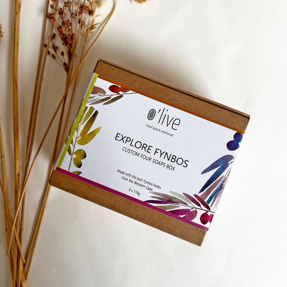 A brown box with a white band that reads 'EXPLORE FYNBOS FOUR SOAPS BOX' presented next to dried grasses. 