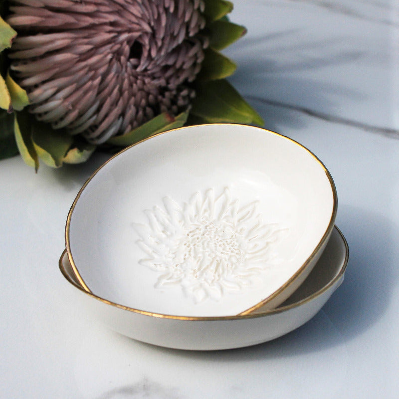 Two white ceramic dipping bowls stacked on top of each other with a gold rim, featuring a king protea design, placed on a marble surface with a large flower in the background.