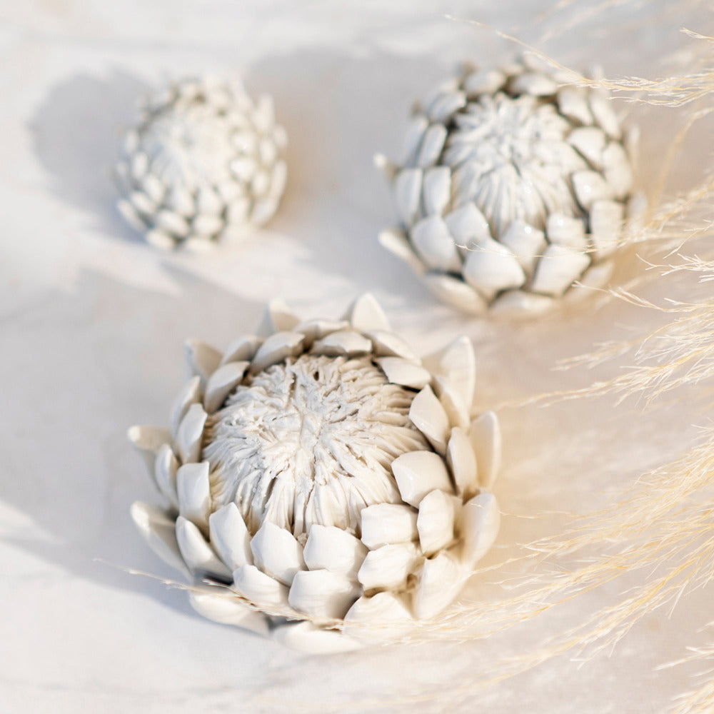 A hand sculpted white porcelain paperweight in the shape of a King Protea flower, displayed on a textured surface.