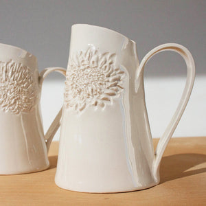 Two white ceramic jugs with protea patterns, placed on a wooden surface against a white background.