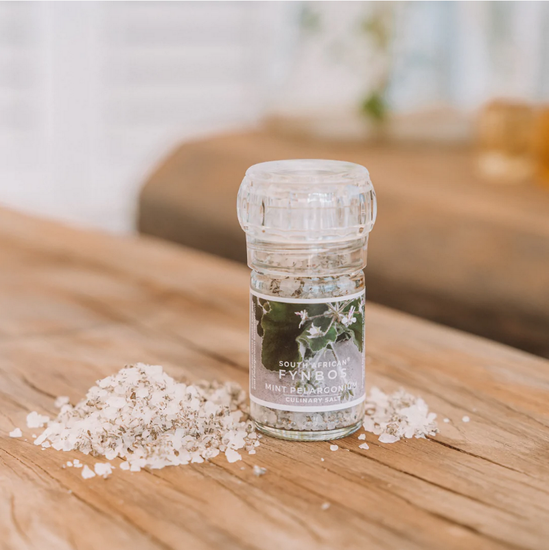 A clear glass culinary grinder labeled 'Mint Pelargonium' with visible white salt crystals next to it on a wooden surface.