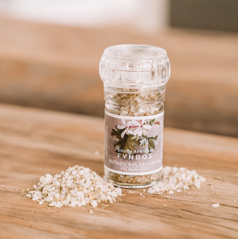 A clear glass culinary grinder labeled 'Nutmeg Pelargonium' with visible white salt crystals next to it on a wooden surface.