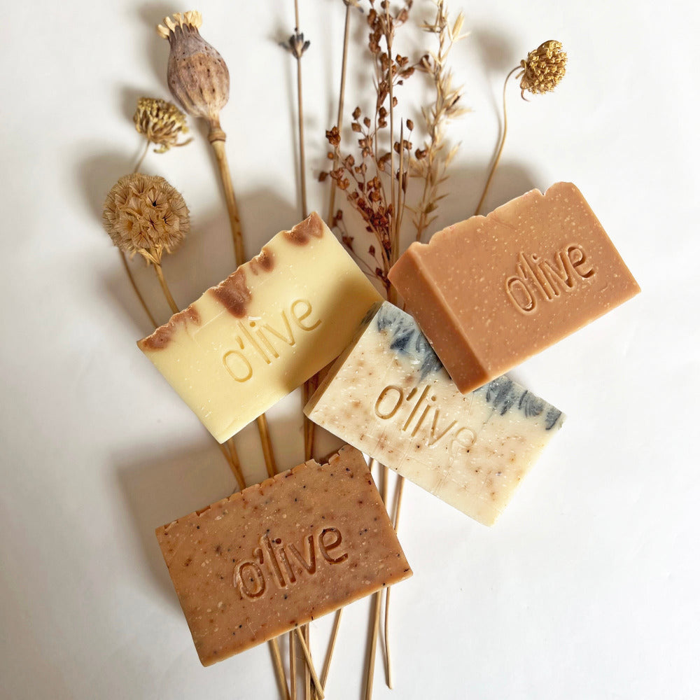 Four handmade soaps engraved with the word 'O'LIVE' displayed on a bed of dried botanicals. 