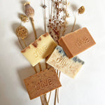 Load image into Gallery viewer, Four handmade soaps engraved with the word &#39;O&#39;LIVE&#39; displayed on a bed of dried botanicals. 
