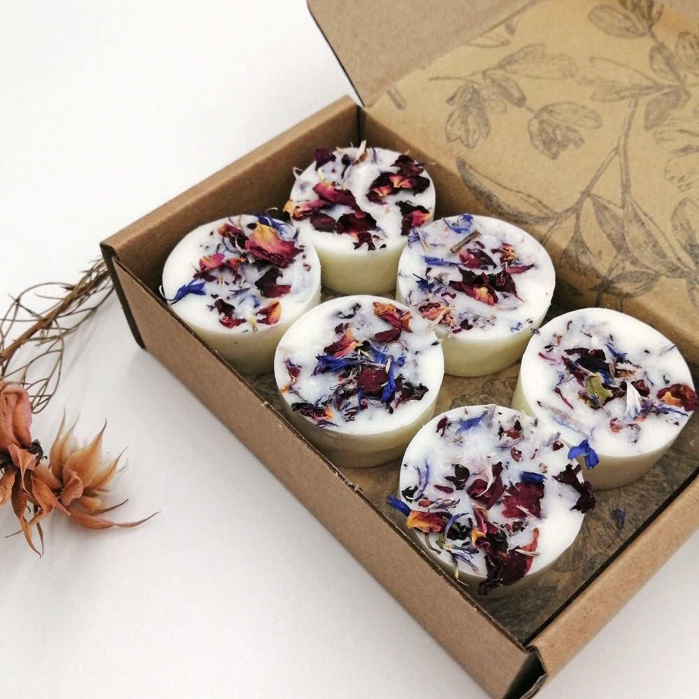 A box of 12 white soy wax melts with multicolored floral toppings, placed in a cardboard box with a floral design. Some dried flowers are also visible to the side.