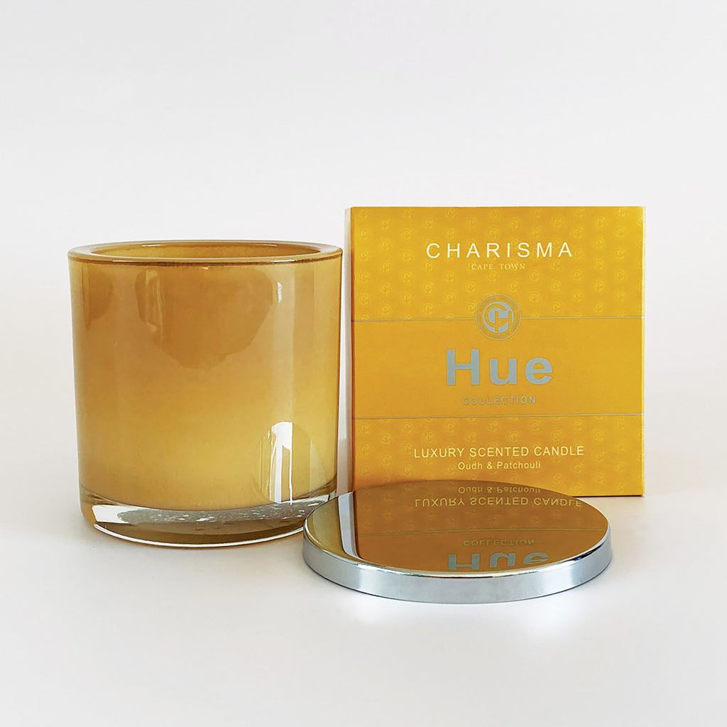 A large single-wick oud and patchouli candle in a tan glass container, with the brand 'Charisma' visible on the packaging and the product name 'HUE' on the candle. The packaging is yellow with the brand logo and product details.