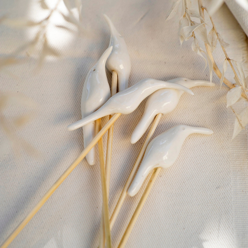 Hand sculpted porcelain sunbird diffuser sticks mounted on bamboo, displayed on a white textured background with dried plant reeds in the peripheral.