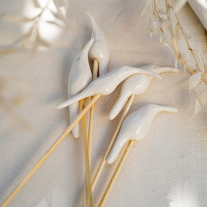 Hand sculpted porcelain sunbird diffuser sticks mounted on bamboo, displayed on a white textured background with dried plant reeds in the peripheral.