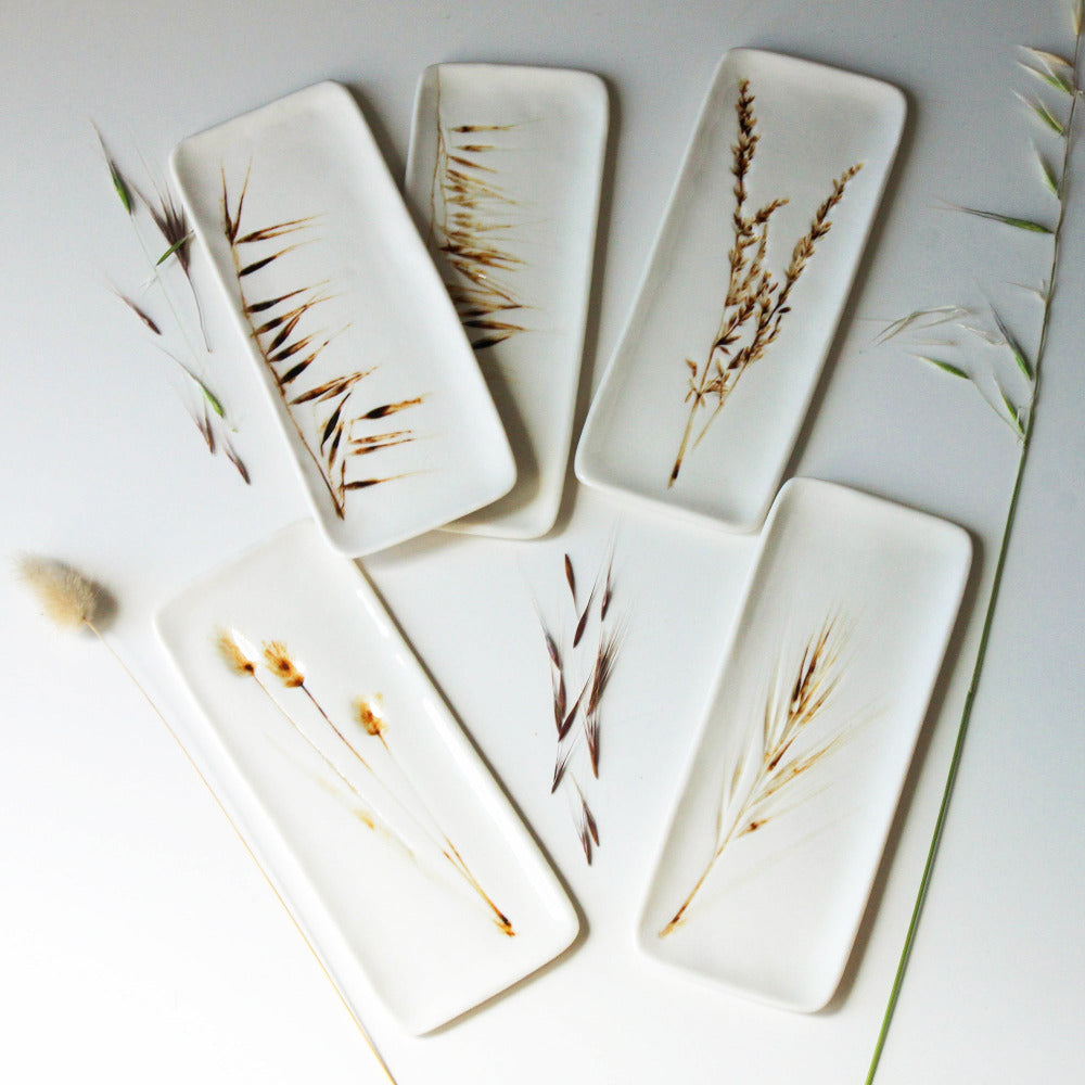 Five white ceramic slates with various botanical designs, including grasses and restio fynbos, displayed on a white surface with a few dried plant accents.