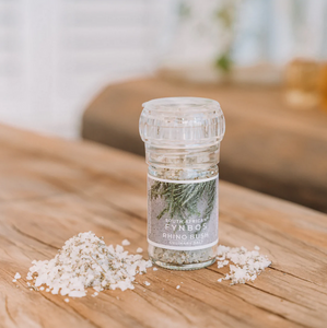 A clear glass culinary grinder labeled 'Rhino Bush' with visible white salt crystals next to it on a wooden surface.