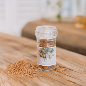 A clear glass culinary grinder labeled 'Rooibos' with visible salt crystals next to it on a wooden surface.