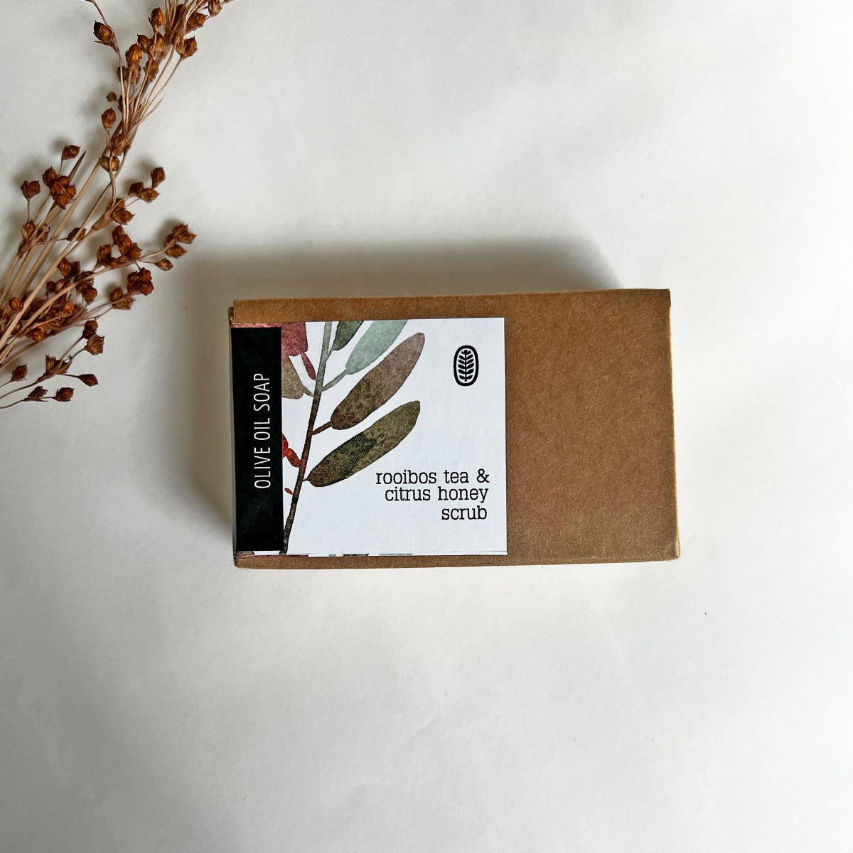 A bar of olive oil soap with rooibos tea & citrus honey scrub, accompanied by dried grass, placed on a white background.