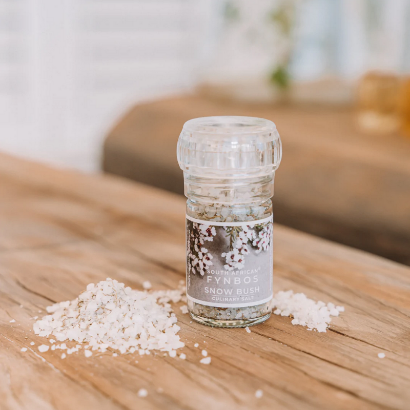 A clear glass culinary grinder labeled 'Snow Bush' with visible white salt crystals next to it on a wooden surface.