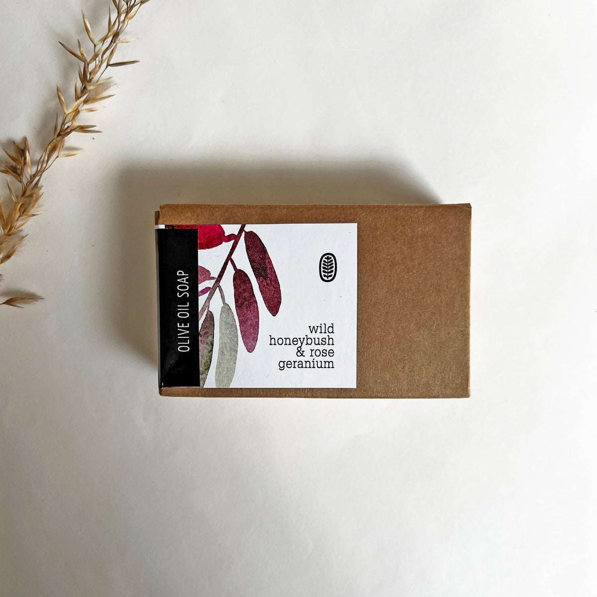 A bar of olive oil soap with wild honeybush & rose geranium, accompanied by dried grass, placed on a white background.