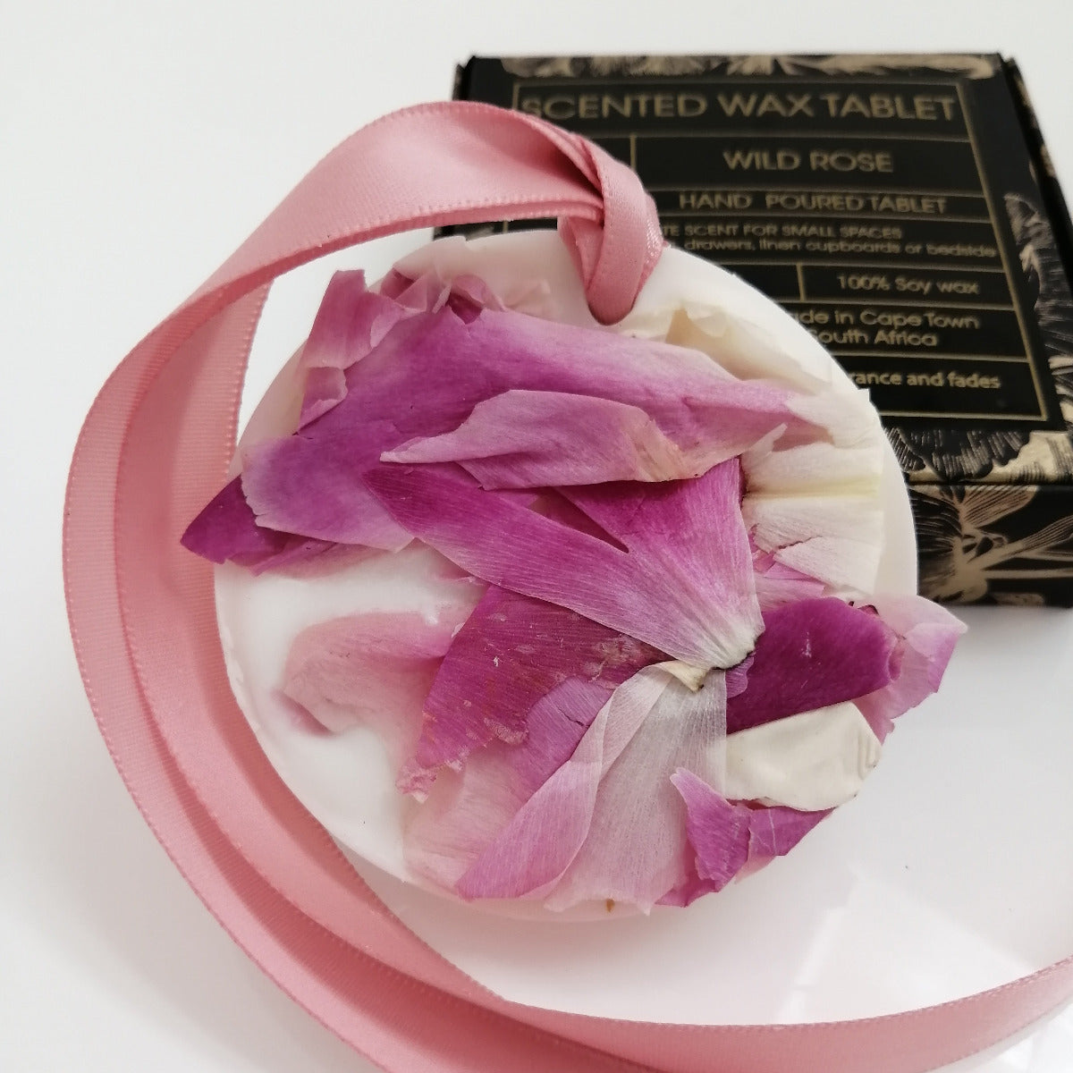 Decorative wax tablet with pink and white petals and a pink ribbon, with a box labeled 'Scented Wax Tablet Wild Rose' in the background.
